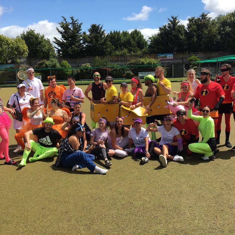 A large group of participants at a themed event on the bowling green, wearing colourful costumes and posing for a group photo with smiles and playful expressions.