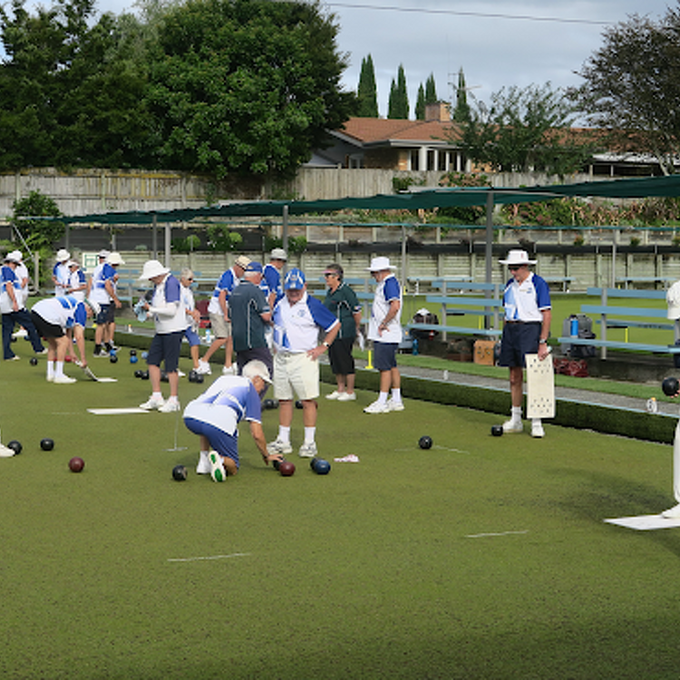Claudelands Bowling Club members engaged in a game on the green, wearing uniforms and hats, with several players observing and a few preparing to bowl.
