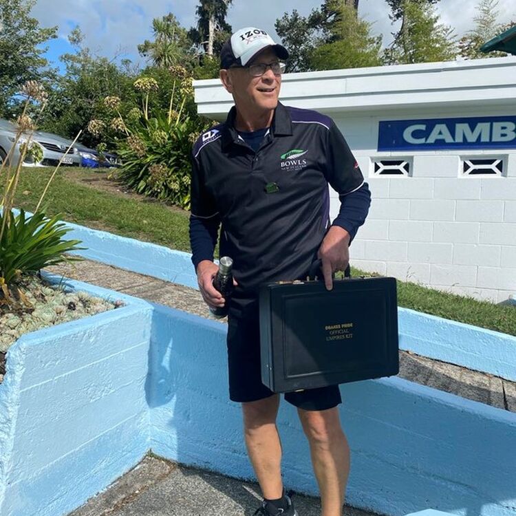 A male club member in a navy blue uniform standing outdoors at the club grounds, holding a black case labeled “Bowls Umpire Official Kit,” with the club sign visible in the background.