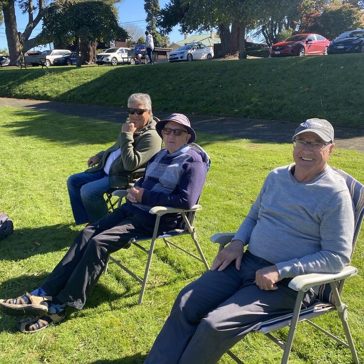 Three spectators seated comfortably in lawn chairs by the green, watching the game with relaxed expressions on a sunny day.