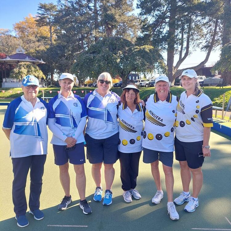 A group of six female club members from Claudelands Bowling Club and Hinuera Club standing together on the green, smiling, dressed in respective club uniforms.