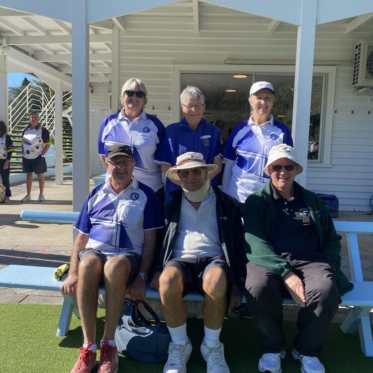 Six Claudelands Bowling Club members seated and standing together on a bench under a shaded area, smiling and enjoying the club environment.
