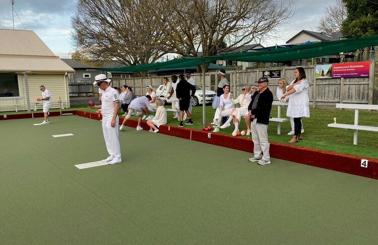 Club members in traditional white uniforms gathered around the bowling green, some seated and some standing, watching a game in progress on an overcast day.