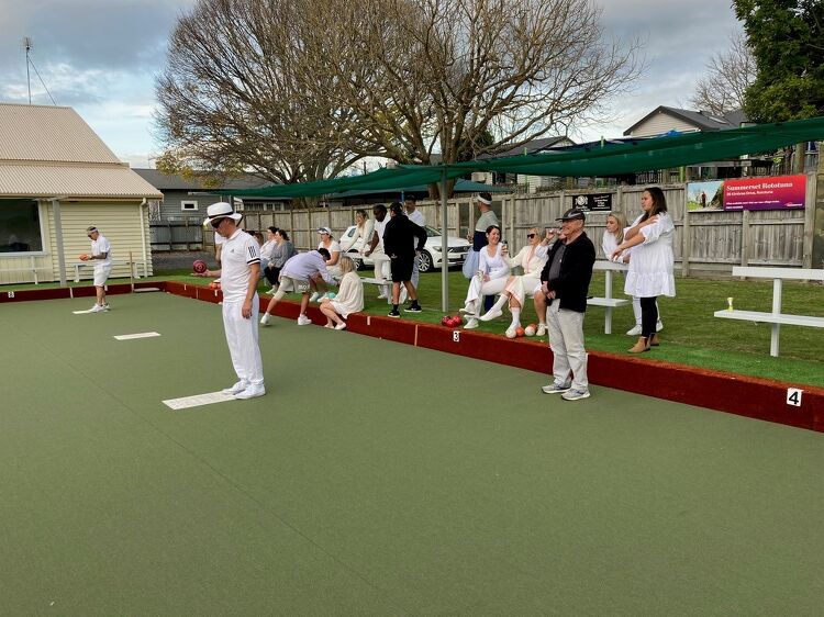 Club members in traditional white uniforms gathered around the bowling green, some seated and some standing, watching a game in progress on an overcast day.