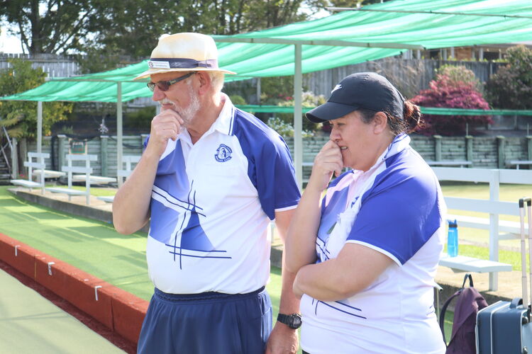 Two members of the Claudelands Bowling Club in white and blue club uniforms, standing on the green, thoughtfully observing the game in progress.