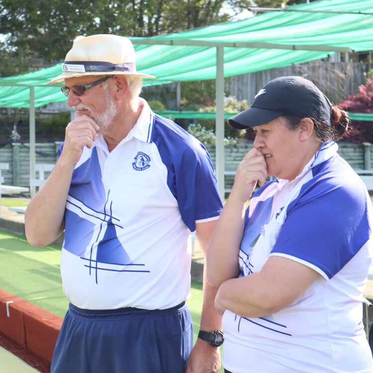 Two members of the Claudelands Bowling Club in white and blue club uniforms, standing on the green, thoughtfully observing the game in progress.