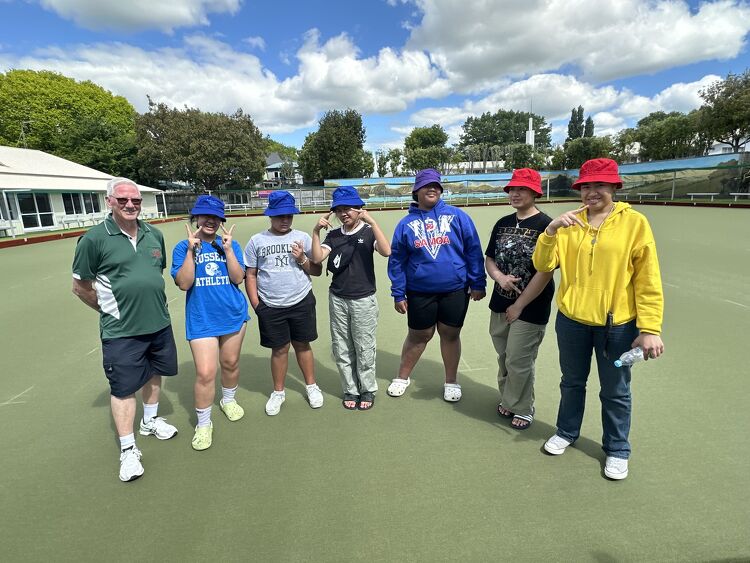 A group of young players wearing colorful bucket hats, along with a Claudelands Bowling Club member in a green shirt, posing together on the bowling green.